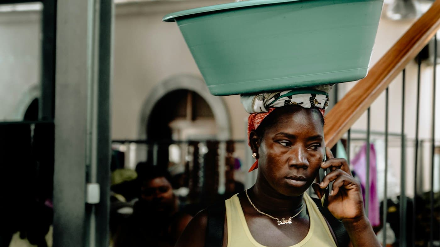 A woman carrying a basin on her head takes a phone call in the street