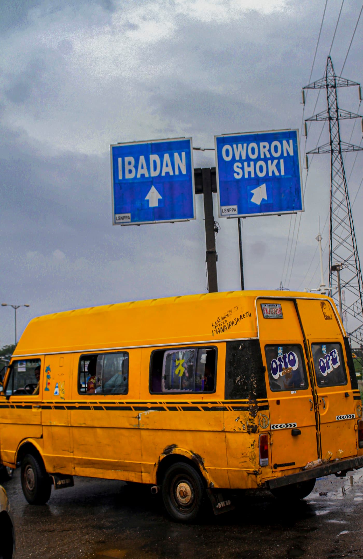 A yellow Lagos danfo bus under road signs for Ibadan and Oworonshoki